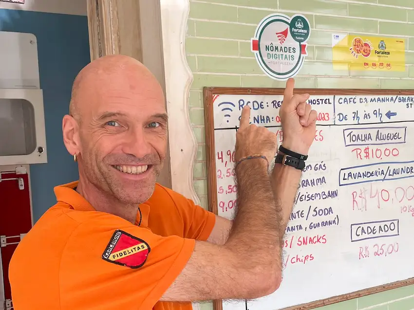 A mostra o Karl, dono do Refúgio Hostel Fortaleza e Pousada apontando sobre o selo Nômades Digitais Fortaleza que foi entregue em junho 2024