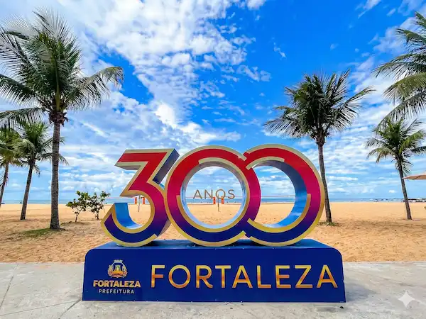 Letreiro turístico em 3D com os números "300" e a palavra "FORTALEZA" em azul, vermelho e dourado, sobre uma base com a logo da Prefeitura. O monumento está no calçadão da Praia de Iracema, cercado por areia, coqueiros e com o mar e um céu ensolarado ao fundo. Ponto turístico próximo ao Refúgio Hostel Fortaleza e à Refúgio Pousada Fortaleza.
